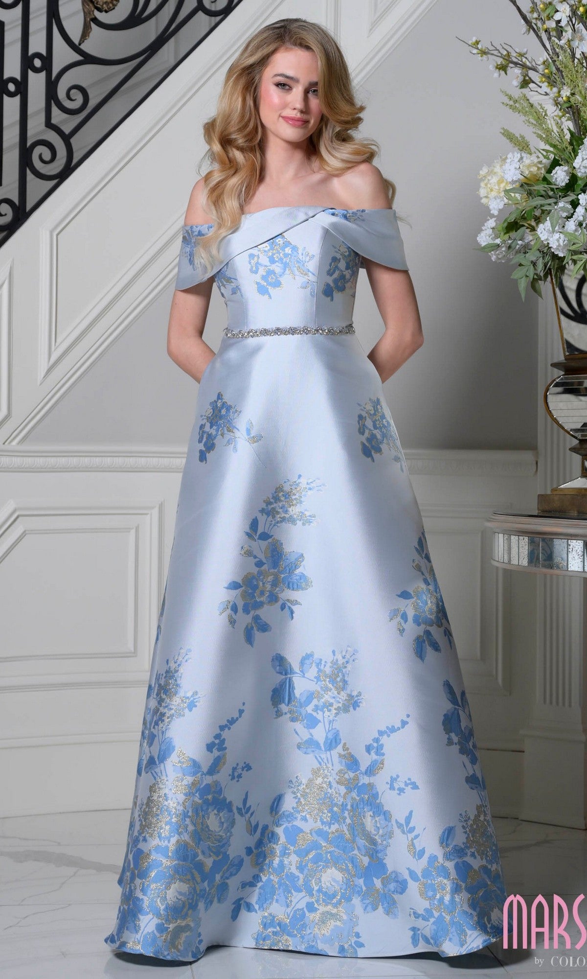 A woman stands indoors wearing the Marsoni MV1402 long formal dress in Blue Multi, featuring jacquard fabric, blue and silver floral patterns, an off-the-shoulder neckline, and a beaded belt.